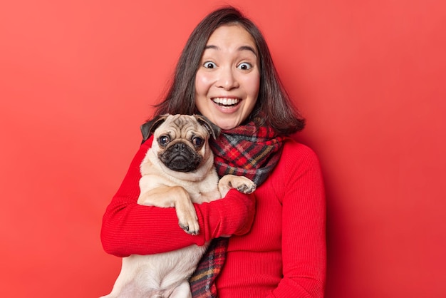 Photo of cute brunette young Asian woman looks with glad surprised face expression smiles happily poses with pug dog going to visit vet clinic dressed casually isolated over vivid red background.