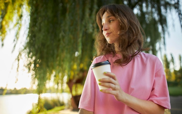 ピンクの t シャツを着たかわいい魅力的な若い女性の写真、公園で、紙のコーヒー カップを持って笑顔で。