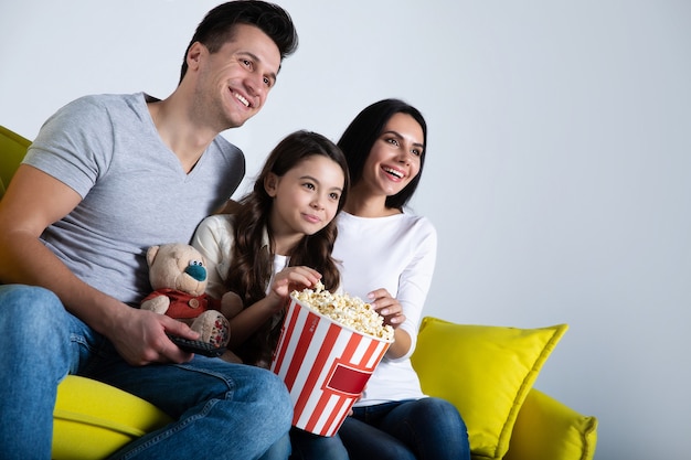 Photo of a cute child and her parents, who are eating popcorn together, while watching TV shows on the couch.