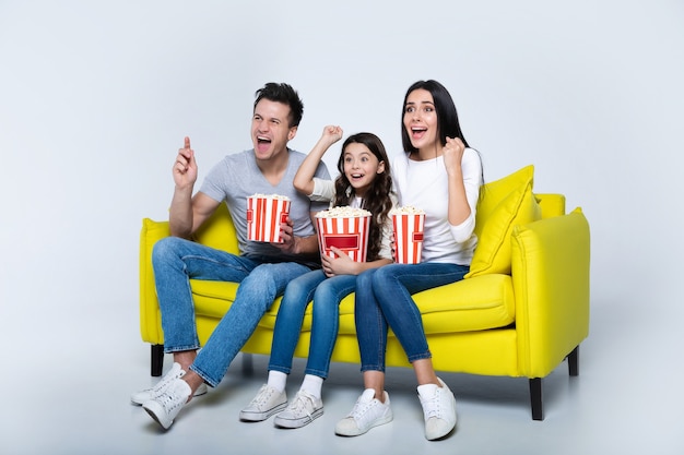 Photo of a cute child and her parents, who are eating popcorn together, while watching TV shows on the couch.