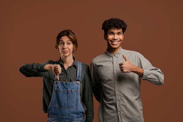 Photo of cute couple farmers. Female farmer shows thumb down but male shows up. Woman wears denim overalls, man wears t-shirt, isolated brown color background.