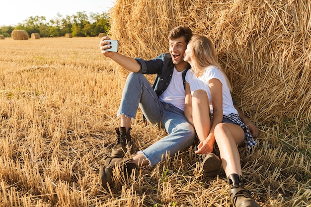 Foto di coppia carina uomo e donna prendendo selfie mentre è seduto sotto un grande pagliaio in campo dorato, durante la giornata di sole