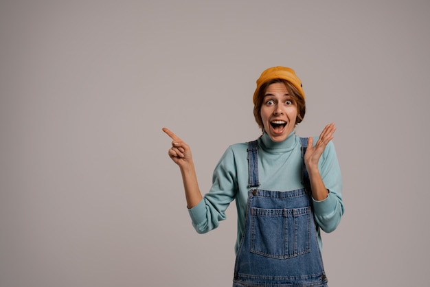 Photo of cute female hipster unreally shocked and points at left side. White woman wears denim overall and hat isolated grey color background.