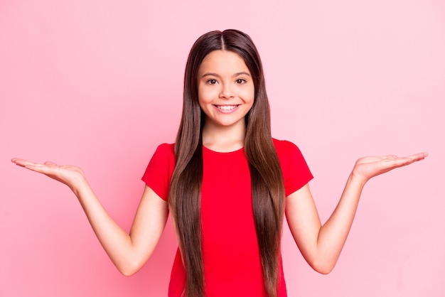 Photo of cute sweet cheerful little lady long hairstyle two open palms arms holding empty space toothy shiny smiling positive face look wear red t-shirt isolated pink color background