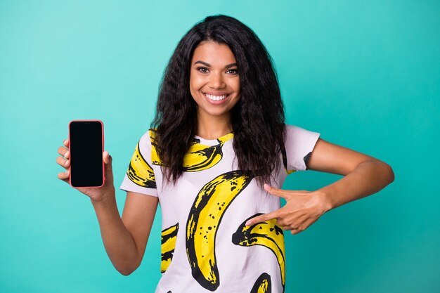 Photo of cute young girl hold smartphone screen empty space direct finger wear banana print t-shirt isolated teal color background