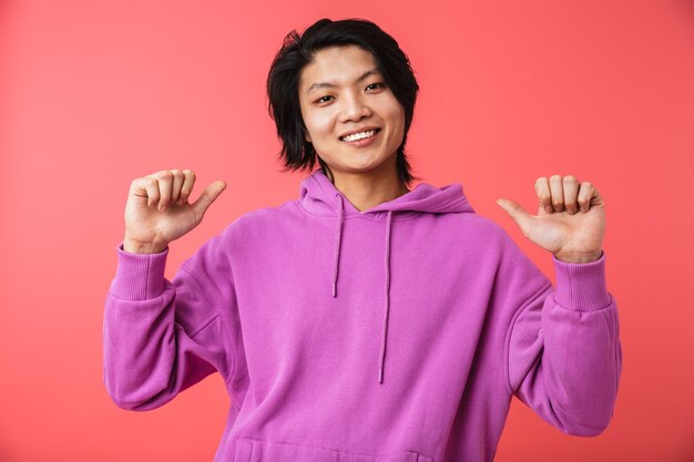 Photo of delighted asian guy wearing sweatshirt rejoicing and pointing fingers at himself isolated over red wall