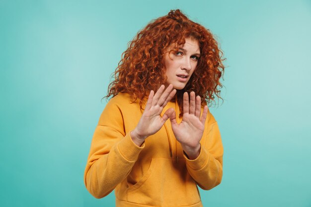 Photo of displeased woman 20s with curly ginger hair doing stop gesture and expressing rejection isolated over blue wall