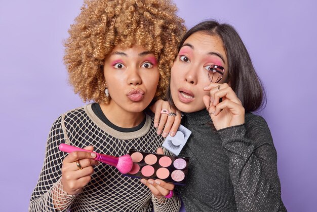 Photo of diverse young women use eyelashes curler and cosmetic\
brush hold colorful eyeshadow palette dressed in casual jumpers\
look impressed isolated over purple background makeup concept