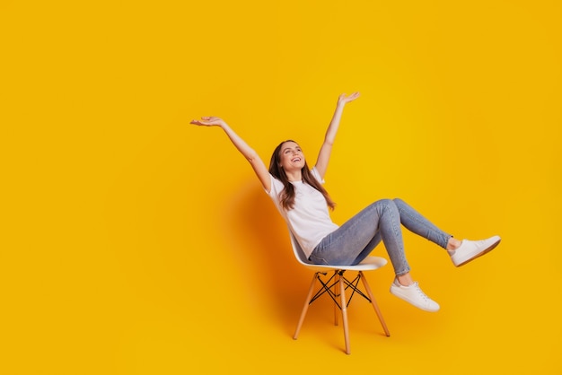 Foto di una donna ispirata ai sogni si siede sedia alza le braccia cerca lo spazio vuoto indossa una t-shirt bianca in posa su sfondo giallo