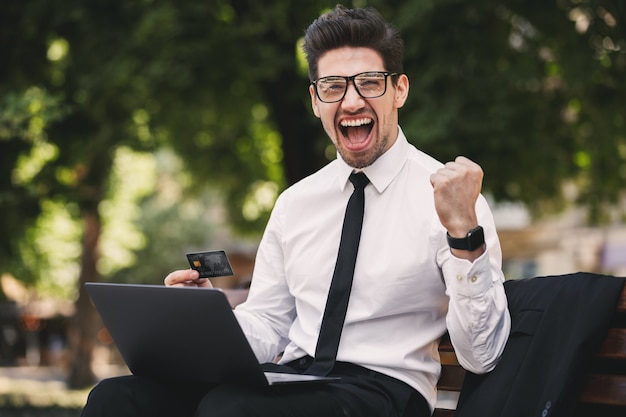 Photo of ecstatic businessman in suit screaming while sitting on bench in green park, and working on laptop during sunny day