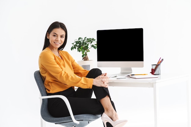 Photo of elegant brunette businesswoman 20s sitting at table and working on big computer in bright office