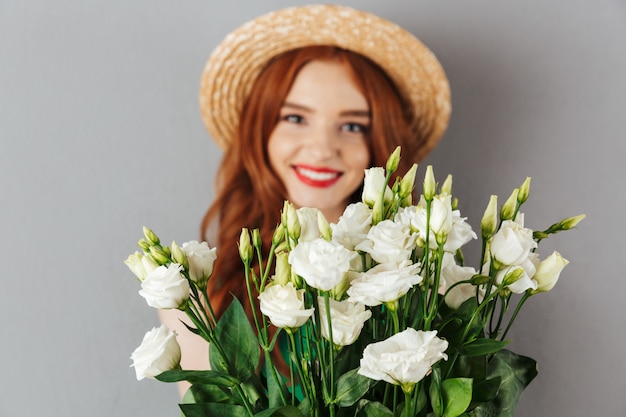 Foto della donna elegante 20s con capelli rossi che indossa il cappello di paglia e che tiene mazzo di eustoma dei fiori bianchi, isolato sopra la parete grigia