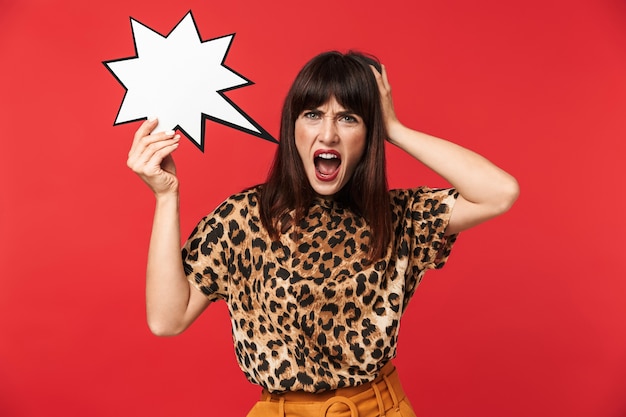 Photo of emotional brunette woman 30s dressed in stylish outfit screaming and holding blank speech bubble isolated over red wall