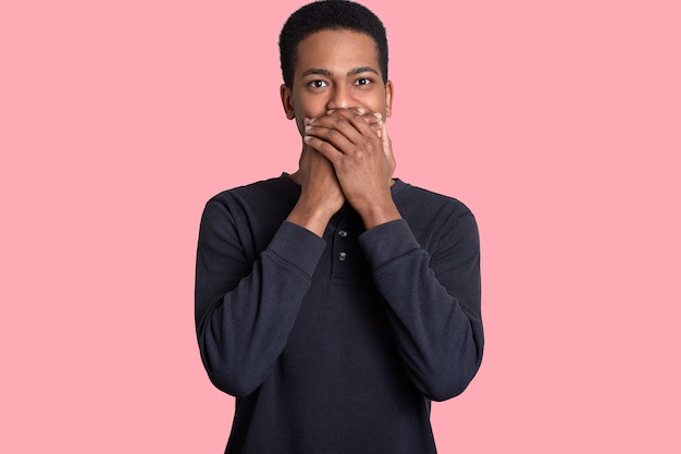 Photo of emotional dark skinned man cons mouth with both palms, dressed in casual jumper, poses on pink, cant stop laughing or being speechless. Ethnicity and gesturing concept