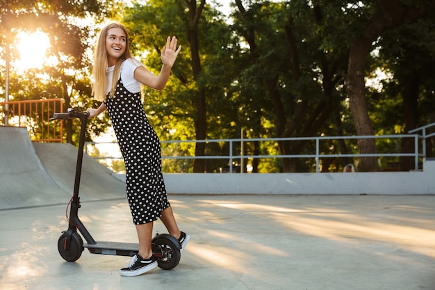 Photo of emotional optimistic cheerful teenage girl in park walking on scooter waving to friends.