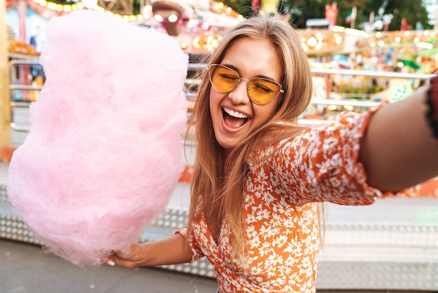 Photo photo of emotional positive cute woman walking outdoors in amusement park eat candyfloss take selfie by camera.