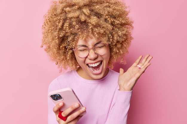 Photo of emotional woman with blonde curly hair exclaims loudly keeps palm raised uses mobile phone gets messages from boyfriend and friends wears transparent glasses jumper isolated on pink wall