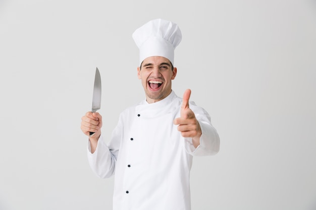 Photo of emotional young man chef indoors isolated over white wall background holding knife pointing.