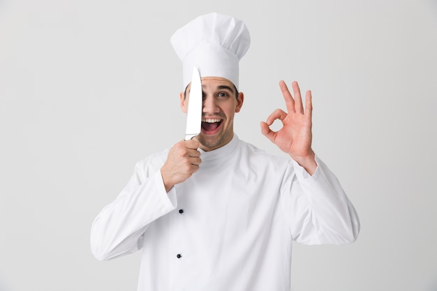 Photo of emotional young man chef indoors isolated over white wall background holding knife showing okay gesture.