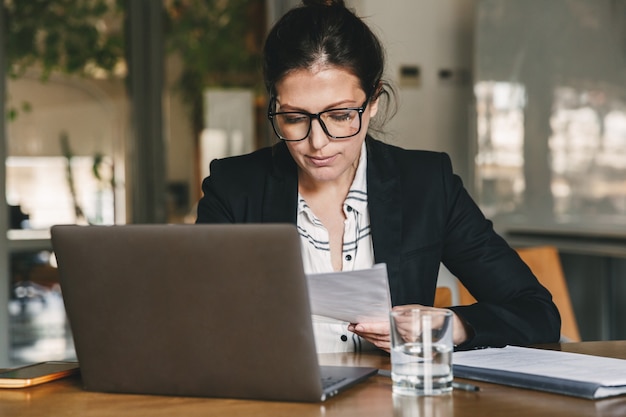 Photo of european businesswoman 30s wearing formal clothing and eyeglasses working in office on laptop and examining paper documents, while holding in hand