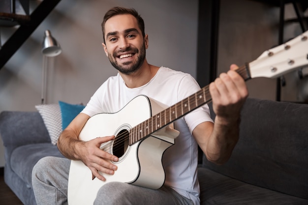 Photo of european man 30s wearing casual t-shirt playing acoustic guitar while sitting on sofa in apartment