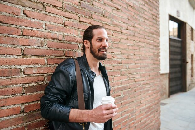 Foto di un uomo non rasato europeo con i capelli legati in piedi sopra il muro di mattoni sulla strada della città e bere caffè da asporto