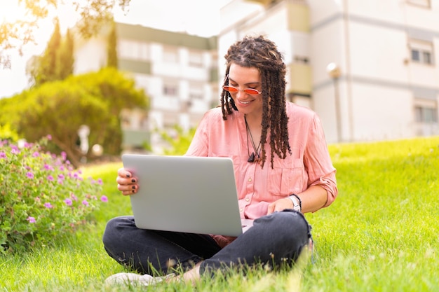 Photo of european woman 25s sitting on green grass in park with legs crossed during summer day while using laptop Caucasian female hipster with dreadlocks and pink sunglasses on use laptop