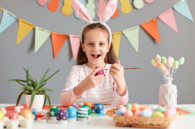 Photo of excited adorable little girl painting aster eggs with brush wearing bunny ears looking at camera with happiness celebrating Easter posing against decorated gray background