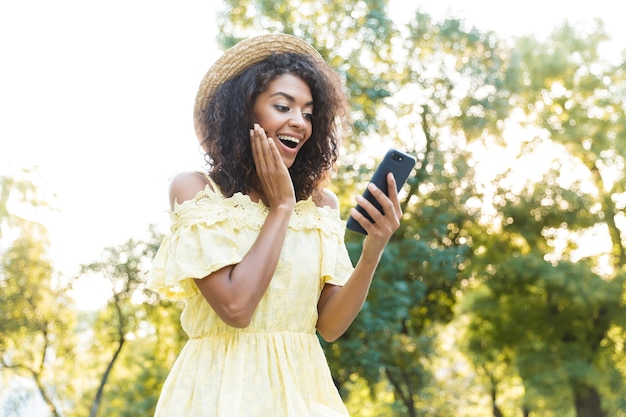 公園を歩きながらスマートフォンを使用して、麦わら帽子をかぶった20代の興奮したアメリカ人女性の写真