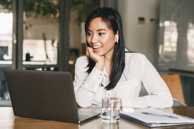 Photo photo of excited asian woman 20s wearing white shirt and bluetooth earphones smiling, while looking on laptop sitting at table in office