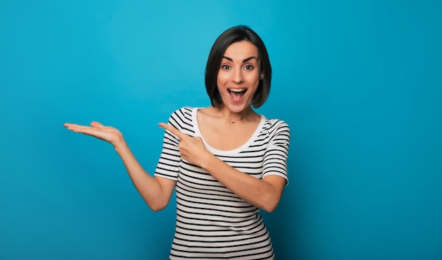 Foto foto di una donna bruna attraente eccitata in abbigliamento casual mentre indica qualcosa di pubblicitario sul braccio e isolata su sfondo blu