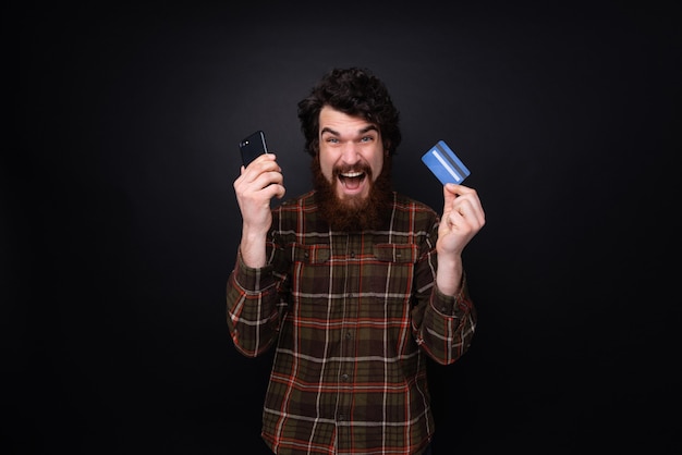 Photo of excited bearded guy with credit card and smartphone in hands standing over dark background