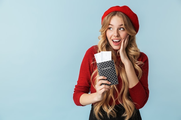 Photo of excited blond girl 20s wearing red beret holding passport and travel ticket isolated 