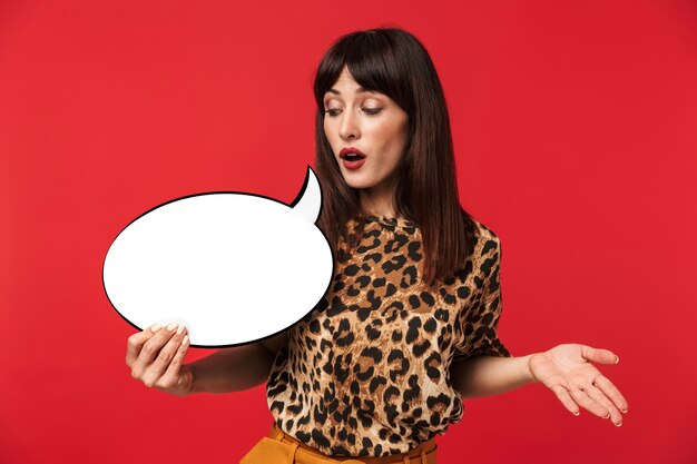 Photo of excited brunette woman 30s dressed in stylish outfit smiling and holding blank thought bubble isolated over red wall