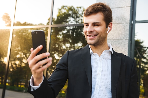 Photo of excited businessman in suit holding mobile phone, while standing outdoor against building