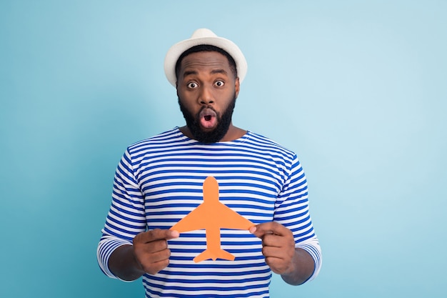 Photo of excited dark skin guy traveler hold paper air plane open mouth win lottery free trip abroad resort wear white sun cap striped sailor shirt isolated blue color wall