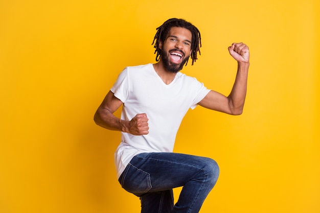 Photo of excited dark skin model guy raise fists leg celebrate goal match wear casual t-shirt isolated yellow color background