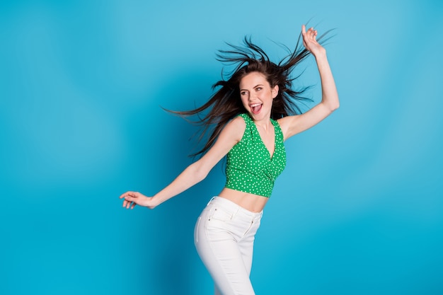 Photo of excited energetic girl dance discotheque air wind blow hair raise hands wear tank-top trousers isolated over blue color background