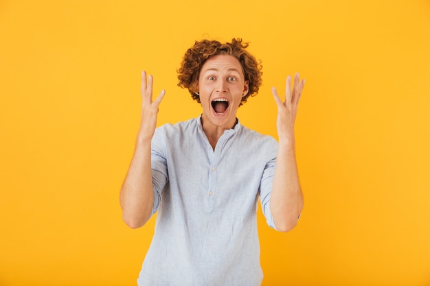 Photo of excited european man  shouting and raising arms in surprise, isolated over yellow background