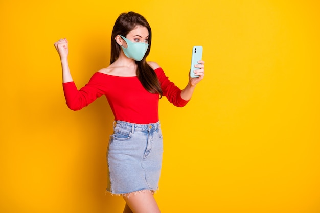 Photo of excited girl in medical mask use smartphone raise fists read social media covid victory news wear red top short mini skirt isolated over bright shine color background