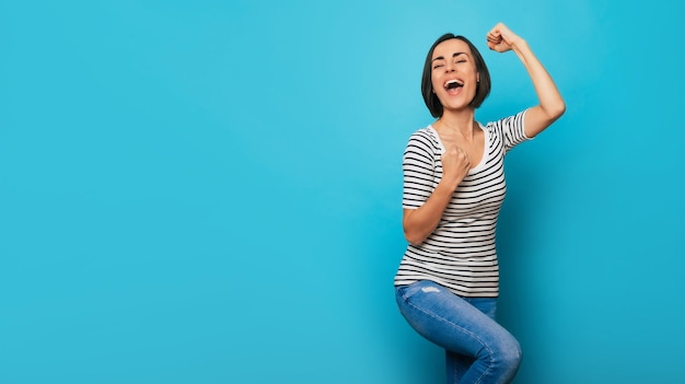 Photo of an excited and happy beautiful young woman with fists up when she shouting and celebrating some win or good news isolated on blue background