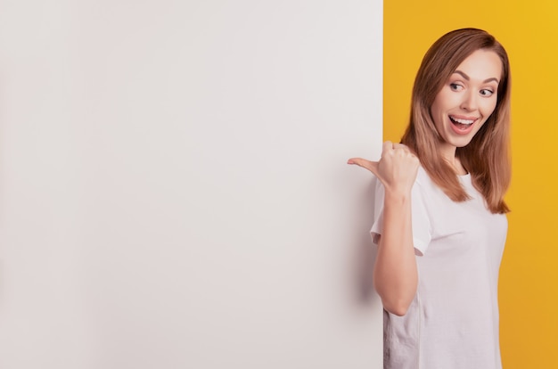 興奮した女性の写真は、黄色の壁に親指の空きスペースが口を開いていることを示しています
