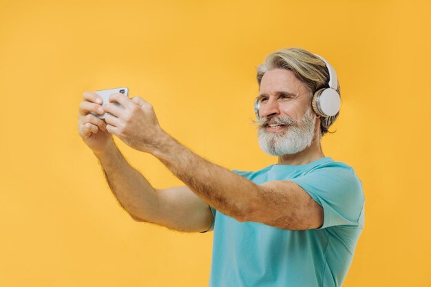 Photo of an expressive grayhaired senior man in headphones with a phone in his hands isolated on a yellow background