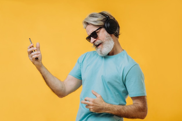 Photo of an expressive grayhaired senior man in headphones with\
a phone in his hands isolated on a yellow background