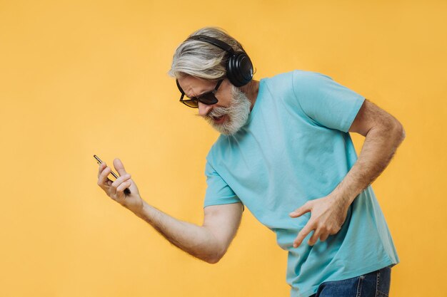 Photo of an expressive grayhaired senior man in headphones with\
a phone in his hands isolated on a yellow background