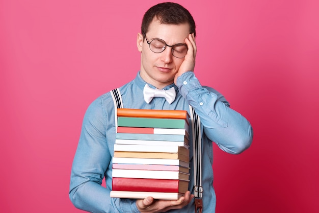 Photo of exuasted handsome student, holding huge stack of books, suffers from terrible headache, posing with hand on temple