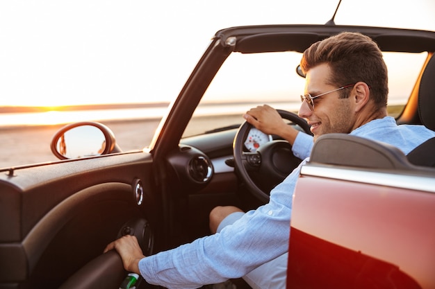 Photo of fashionable caucasian man wearing sunglasses getting out of convertible stylish car by seaside at sunrise