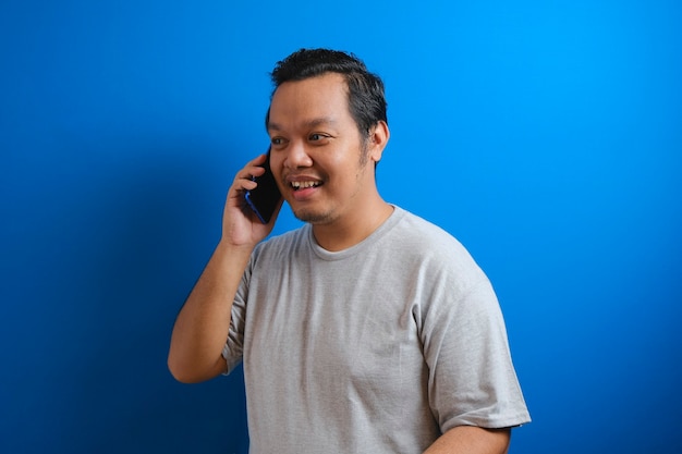 Photo of a fat asian man wearing a gray shirt smiling while receiving a phone. isolated on blue background