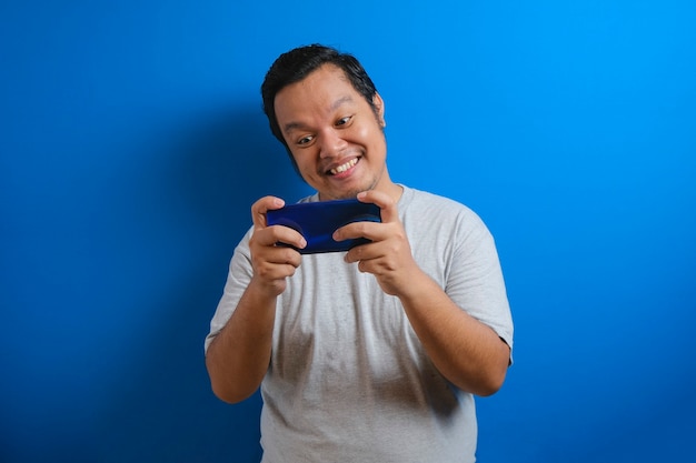 회색 티셔츠를 입은 뚱뚱한 아시아 남성의 사진은 스마트폰으로 게임을 하며 행복해 보입니다. 남자는 자신감 있는 제스처를 보여줍니다. 파란색 배경에 고립