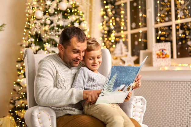 Photo of father and son talk enjoy noel eve, rest and reading miracle story carol, wear sweater in decorated x-mas home indoors